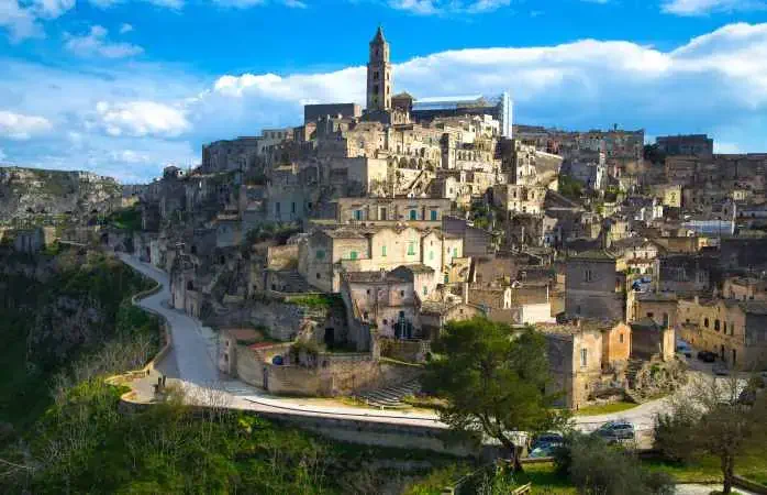 Beautiful cityscape of Matera against the blue sky. Large white clouds in the sky. City in the rock. The winding road around the city. Green grass and trees. Italy.